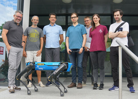 Sieben Personen stehen vor dem Eingangsbereich eines Gebäudes. Im Vordergrund steht ein blauer vierbeiniger Roboter mit schwarzer Struktur – offensichtlich ein Forschungsroboter.