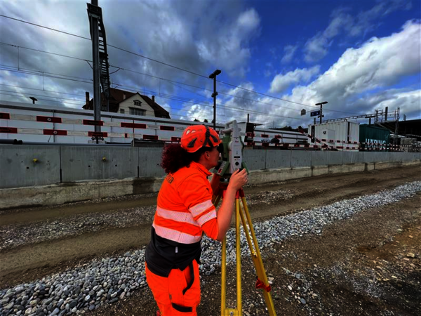 A worker in bright orange high-visibility safety clothing and helmet operates a tripod-mounted surveying instrument beside a railway yard, with tracks, barriers, and a blue sky with clouds in the background.
