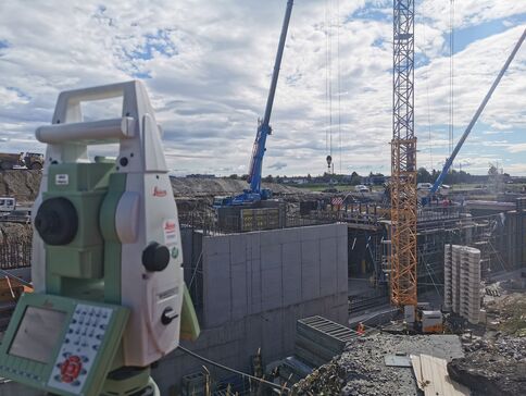 Auf einer Baustelle arbeiten Kräne, Betonwände und Baugerüste; im Vordergrund steht ein Vermessungsgerät zur Bauvermessung.