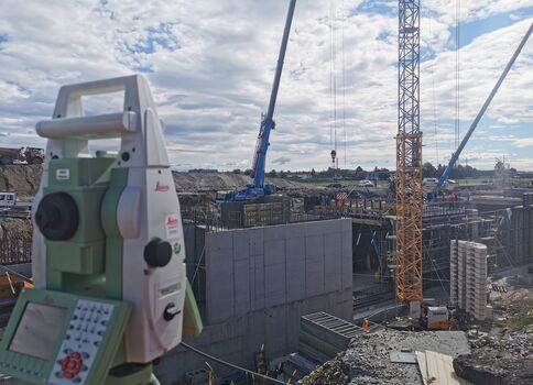 Auf einer Baustelle arbeiten Kräne, Betonwände und Baugerüste; im Vordergrund steht ein Vermessungsgerät zur Bauvermessung.