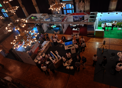 Blick von oben auf eine Messehalle mit mehreren Messeständen, Monitoren und Besuchern im Gespräch. Kronleuchter hängen von der Decke; rechts ein grüner Ausstellungsbereich mit Stühlen.