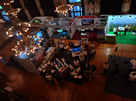 Blick von oben auf eine Messehalle mit mehreren Messeständen, Monitoren und Besuchern im Gespräch. Kronleuchter hängen von der Decke; rechts ein grüner Ausstellungsbereich mit Stühlen.