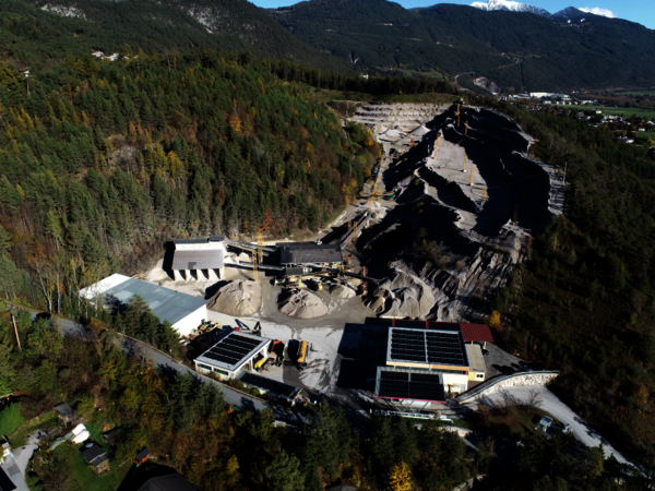 Luftaufnahme eines offenen Bergbaus in einer bewaldeten Gebirgslandschaft. Ein großes, gestuftes Abbaugebiet aus dunklem Material mit Gebäuden, Lagerflächen und Straßen; umliegende Wälder und Bergketten im Hintergrund.