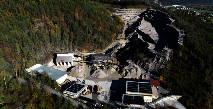 Luftaufnahme eines offenen Bergbaus in einer bewaldeten Gebirgslandschaft. Ein großes, gestuftes Abbaugebiet aus dunklem Material mit Gebäuden, Lagerflächen und Straßen; umliegende Wälder und Bergketten im Hintergrund.
