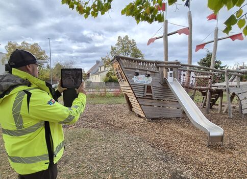 Eine Person in fluoreszierend gelber Warnjacke filmt oder fotografiert ein hölzernes Kletter- und Spielgerät mit Rutsche auf einem Spielplatz; im Hintergrund stehen Bäume, der Himmel ist wolkig.