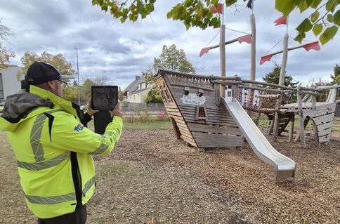 Eine Person in fluoreszierend gelber Warnjacke filmt oder fotografiert ein hölzernes Kletter- und Spielgerät mit Rutsche auf einem Spielplatz; im Hintergrund stehen Bäume, der Himmel ist wolkig.