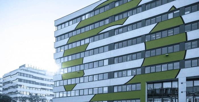 Mehrstöckiges, modernes Gebäude mit auffälliger Fassadengestaltung: weiße und graue Flächen sowie grüne Kreise, die Muster über die Fassade bilden; im Hintergrund weitere Gebäude.