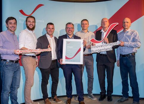 Sieben Männer posieren auf einer Bühne während einer Preisverleihung. In der Mitte hält einer eine gerahmte Urkunde; links und rechts tragen weitere zwei Personen Schilder mit Hashtags, und rechts befindet sich ein Schild mit "Innovationspreis Burgenland". Der Hintergrund ist ein blaues Bühnenbanner.