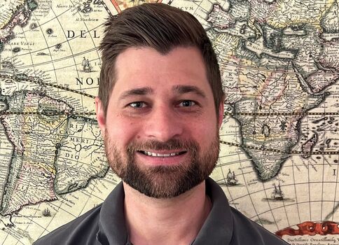 Porträt eines lächelnden Mannes mit Bart vor einer historischen Weltkarte. Er trägt ein dunkles Polohemd mit LG-Logo und blickt in die Kamera.