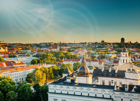 Panorama einer historischen europäischen Stadt. Im Vordergrund ein weißer Gebäudekomplex mit Kuppeln, dahinter Kirchtürme, rote Dächer und grüne Bäume. Weite Stadtlandschaft unter blauem Himmel mit Sonnenlicht.