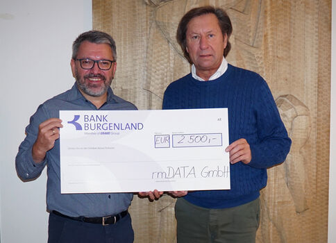 Zwei Männer stehen nebeneinander und halten einen großen Scheck der Bank Burgenland über 2.500 EUR, adressiert an rmDATA GmbH. Hintergrund: beige Vorhang.