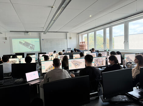 Eine Computerklasse oder CAD-Labor; Studierende sitzen an Tischen mit Desktop- und Laptop-Computern und arbeiten an CAD- oder 3D-Modellierungsprojekten. Vorne wird eine technische Zeichnung projiziert. Große Fenster an der Seite lassen Tageslicht herein.
