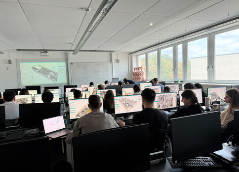 Eine Computerklasse oder CAD-Labor; Studierende sitzen an Tischen mit Desktop- und Laptop-Computern und arbeiten an CAD- oder 3D-Modellierungsprojekten. Vorne wird eine technische Zeichnung projiziert. Große Fenster an der Seite lassen Tageslicht herein.