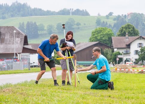 Trois personnes réalisent un relevé topographique en plein air: un homme en bleu tient une perche jaune, une femme consulte un carnet près d'un trépied avec instrument de mesure, et un autre homme en turquoise ajuste l'équipement sur un terrain vert, près de maisons.