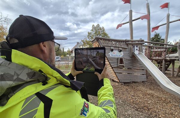 Ein Mann in einer neongelben Sicherheitsjacke und Baseballkappe fotografiert mit einem Tablet einen Spielplatz mit einer Holzrutsche und Klettergerüst; rote Flaggen auf Holzkonstruktionen; der Boden besteht aus Holzspänen; der Himmel ist bewölkt.