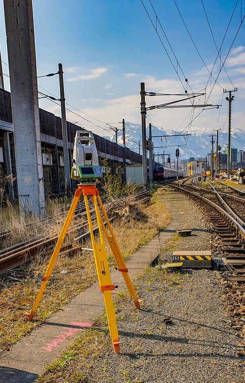 Vermessungsgerät auf einem gelben Dreibein-Stativ steht direkt neben Eisenbahngleisen. Im Hintergrund erkennen Sie Züge, Oberleitungen und weit verlaufende Gleise; am Horizont liegen schneebedeckte Berge.