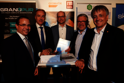 Fünf Männer in Anzügen posieren bei einer Fachmesse. Sie stehen hinter einem Stehtisch mit Unterlagen; einer präsentiert ein Dossier. Im Hintergrund sind Banner mit Logos verschiedener Unternehmen zu sehen.