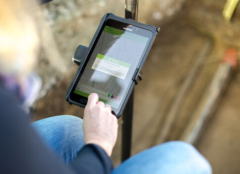 Eine Person sitzt im Freien und bedient ein auf einem Halter montiertes Tablet. Auf dem Display ist eine Vermessungs-/Kartierungs-App zu sehen, offenbar bei einer Bau- bzw. Feldarbeitsituation.