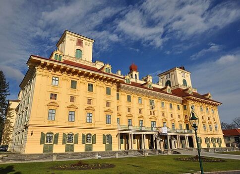 Großes gelb gestrichenes Schloss mit roten Dächern und mehreren Türmen. Die mehrstöckige Fassade zeigt viele Fenster, eine zentrale Eingangstür mit Balkon und bogengeformte Fensterelemente. Vor dem Gebäude eine gepflegte Wiese; der klare blaue Himmel und links einige Bäume runden das Bild.