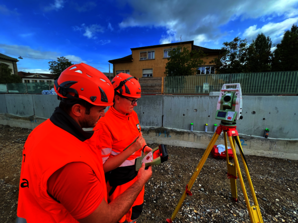 Two construction workers in bright orange safety vests and red helmets operate a tripod-mounted surveying instrument at a construction site, measuring and adjusting equipment against a concrete barrier with a building in the background.