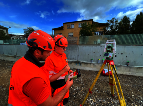 Zwei Bauarbeiter in leuchtend orange Warnkleidung und roten Helmen arbeiten auf einer Baustelle und bedienen ein Messgerät auf einem Stativ; im Hintergrund sind Gebäude und eine Betonmauer sichtbar.