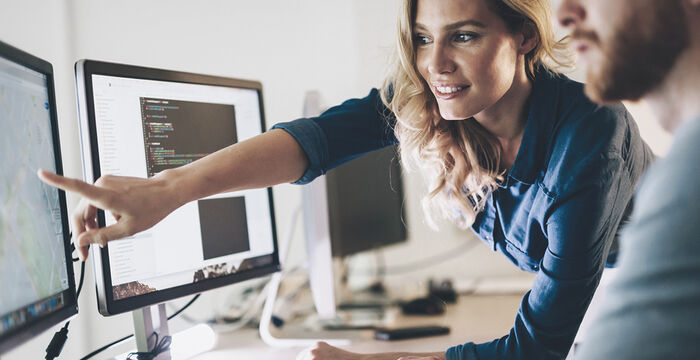 Zwei Softwareentwickler arbeiten gemeinsam an einem Computerarbeitsplatz: Eine Frau zeigt mit dem Finger auf einen Monitor mit Programmiercode, während ein Mann die Maus bedient. Im Hintergrund sind weitere Monitore und Arbeitsmaterialien zu sehen.