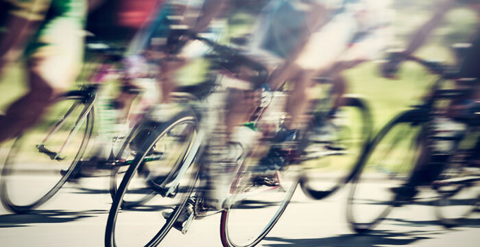 Peloton de cyclistes en course sur une route extérieure; vélos et roues flous par le mouvement, indiquant une compétition sportive dynamique.