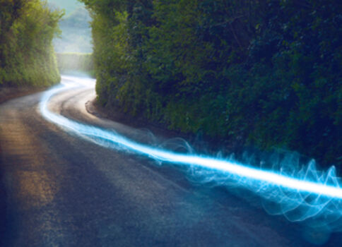 Eine kurvige Straße windet sich durch einen dicht bewaldeten Wald; Bäume säumen beide Seiten, während eine leuchtend blaue Lichtspur sich entlang der Fahrbahn zieht.