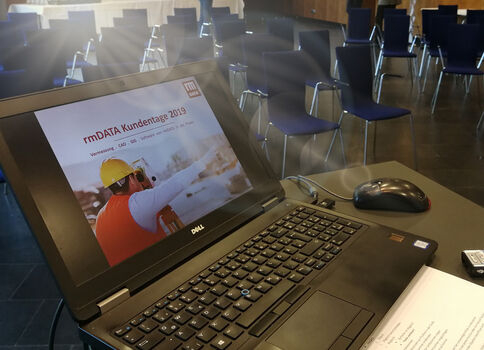 Laptop zeigt eine Präsentationsfolie „rmDATA Kundentage 2019“ mit einem Bauarbeiter, der einen gelben Helm und eine orangefarbene Weste trägt.