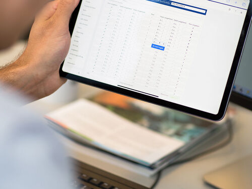 Eine Person hält ein Tablet; auf dem Bildschirm wird eine Website mit einer großen tabellarischen Datentabelle angezeigt, in der Zahlenwerte sichtbar sind.