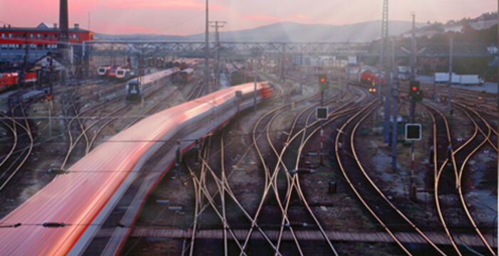 Verschlungene Gleise eines Bahnbetriebswerks; ein roter Hochgeschwindigkeitszug fährt links vorbei, Signale und Oberleitungen überspannen das Netz. Im Hintergrund Gebäude, eine Turmstruktur und ein warmer Abendhimmel.
