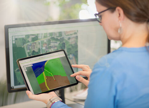 Eine Person in blauer Arbeitskleidung hält ein Tablet mit einer farbigen 3D-Darstellung, deren Flächen grün, blau und braun sind, und tippt darauf; im Hintergrund zeigt ein Monitor eine Kartenansicht.