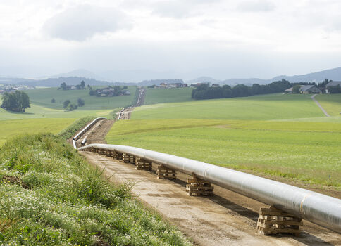 Ländliche Landschaft mit einer langen Rohrleitung, die auf Stützen über einen Feldweg führt. Grüne Felder, sanfte Hügel, entfernte Häuser und ein wolkiger Himmel prägen die Szene.
