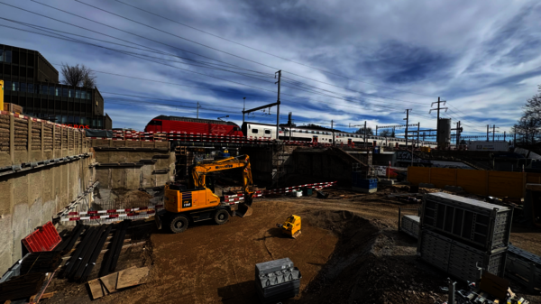 Große Baustelle im Freien: Ein gelb-oranger Bagger und ein kleiner gelber Radlader arbeiten auf aufgeschüttetem Erdreich; Baumaterialien, Stahlreste und Absperrungen liegen um sie herum. Im Hintergrund fährt eine Eisenbahn mit Waggons; der Himmel ist wolkig.