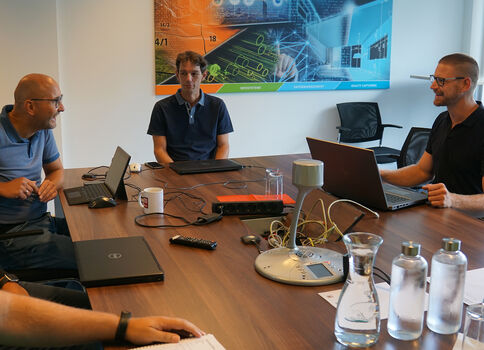 Vier Personen sitzen um einen großen Konferenztisch und arbeiten mit Laptops. Im Hintergrund hängt ein farbiges Poster mit technischen Grafiken. Auf dem Tisch befinden sich Wasserflaschen, Gläser, Kabel und weitere Büroutensilien.