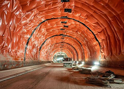 Blick in einen langgestreckten Tunnel im Bau. Die Innenwände sind orangefarben verkleidet, wodurch eine gebogene Tunnelstruktur entsteht. Weiter vorne steht ein Baufahrzeug, am Boden liegen Metall- und Bauholzreste; Arbeitslichter sorgen für Beleuchtung.