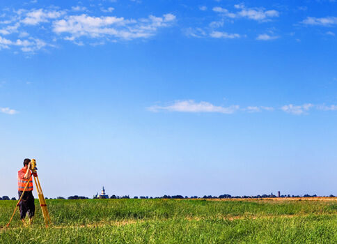 Eine Person steht in einer weiten, grünen Wiesenlandschaft und richtet ein Vermessungsinstrument auf einem Stativ aus; der klare blaue Himmel ist mit wenigen Wolken versehen.