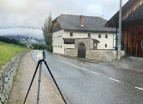 Auf einer ländlichen Straße steht ein Stativ mit Messgerät vor einem weißen, zweigeschossigen Gebäude mit großem Torbogen; links eine Steinmauer, rechts ein weiteres Gebäude. Im Hintergrund liegen Nebel, Wälder und Berge.