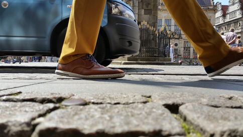 Unterkörper einer Person, die in braunen Schuhen und gelben Hosen über gepflasterten Boden geht; neben ihr steht ein blaues Auto, im Hintergrund eine historische Straßenszene mit Gebäuden.
