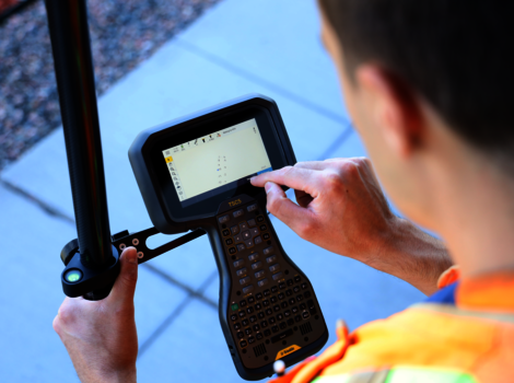 Eine Person in einer leuchtenden Sicherheitsweste bedient ein tragbares Datenterminal mit Tastatur und Touchscreen; eine Hand hält das Gerät, die andere tippt auf dem Bildschirm.