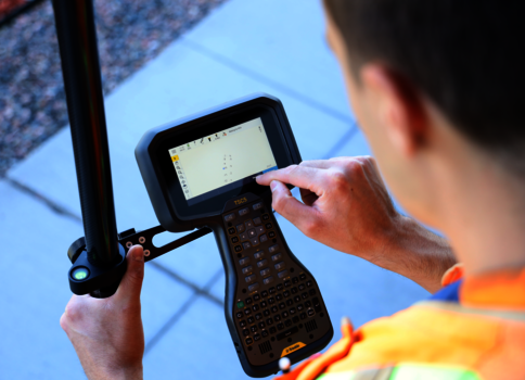 Eine Person in einer leuchtenden Sicherheitsweste bedient ein tragbares Datenterminal mit Tastatur und Touchscreen; eine Hand hält das Gerät, die andere tippt auf dem Bildschirm.