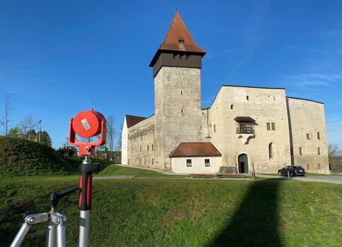 Historische Festung mit einem hohen, steinernen Turm und rotem Spitzdach, von dicken Mauern umgeben. Vor der Anlage liegt eine Grasfläche; der Himmel ist blau und klar, ein Torbogen öffnet den Innenhof.