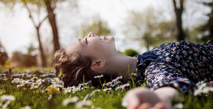 Eine Frau liegt lachend auf einer Blumenwiese, von Sonnenlicht umgeben. Sie trägt ein blumiges Kleid und streckt einen Arm in Richtung Kamera; um sie herum Gras und kleine weiße Gänseblümchen, im Hintergrund Bäume.