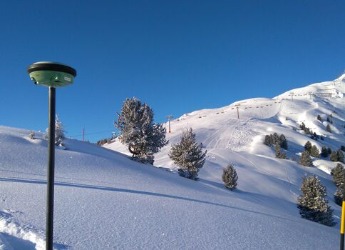 Winterlandschaft: blauer Himmel, schneebedeckte Berge und vereinzelte Tannen. Links oben steht eine Person in grüner Kleidung am Hang; im Vordergrund ein dunkler Laternenpfosten mit rundem Oberteil.