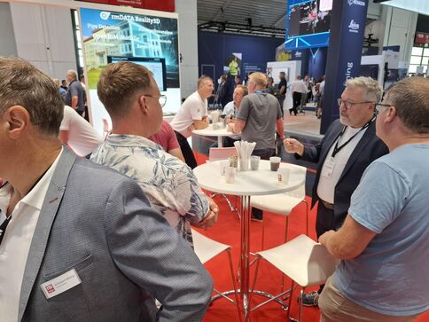 Mehrere Menschen in Business-Kleidung stehen an runden Stehtischen und unterhalten sich in einer Messehalle. Im Hintergrund sind Ausstellerstände, Banner und Bildschirme zu sehen; einige tragen Namensschilder.