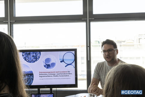 Ein Mann präsentiert in einem Besprechungsraum eine Folie zu Datenmanagement und Governance; im Vordergrund hören mehrere Teilnehmende zu, während ein Monitor Diagramme mit blauem Design zeigt.