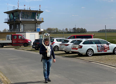 Ein Mann geht auf einer asphaltierten Zufahrt vor einem Kontrollturm auf einem Luftfahrtgelände. Im Hintergrund stehen mehrere geparkte Autos; eine grüne Wiese und Felder erstrecken sich unter klarem Himmel.