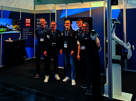 Vier Männer in dunkler Kleidung mit Namensschildern stehen vor einem Tech-Messestand mit blauen Wänden; im Hintergrund befinden sich Bildschirme und ein großer Roboterarm.