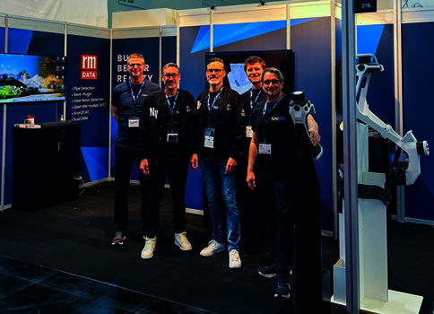 Vier Männer in dunkler Kleidung mit Namensschildern stehen vor einem Tech-Messestand mit blauen Wänden; im Hintergrund befinden sich Bildschirme und ein großer Roboterarm.