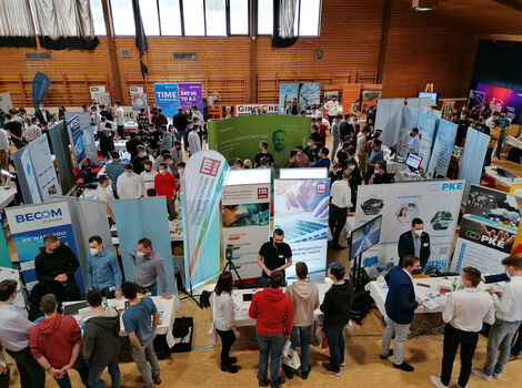 Eine große Turnhalle voller Messestände, Banner und Informationsstände; Besucher erkunden Stände, sprechen mit Ausstellern und sammeln Informationen.